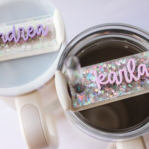 May include: Two white travel mugs with clear lids and pink glitter. The lids have a clear plastic strip with a pink name on each. The mug on the left says "andrea" and the mug on the right says "karla".