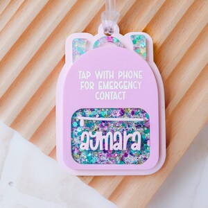 May include: A pink backpack-shaped tag with the text "TAP WITH PHONE FOR EMERGENCY CONTACT" and the name "aymara". The tag has a clear pocket filled with colorful glitter and a clear loop for attaching. The background is a light wood.