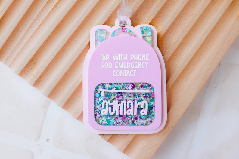 May include: A pink backpack-shaped tag with the text "TAP WITH PHONE FOR EMERGENCY CONTACT" and the name "aymara". The tag has a clear pocket filled with colorful glitter and a clear loop for attaching. The background is a light wood.