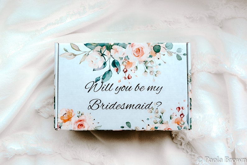 May include: A white box decorated with peach, pink, and green floral patterns, with the words "Will you be my Bridesmaid?" written in script. The box is placed on a soft, white fabric background.