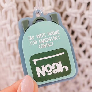 May include: A light blue backpack-shaped tag with the name "Noah" in white. The tag has the text "TAP WITH PHONE FOR EMERGENCY CONTACT". The tag is held up by a clear plastic loop.