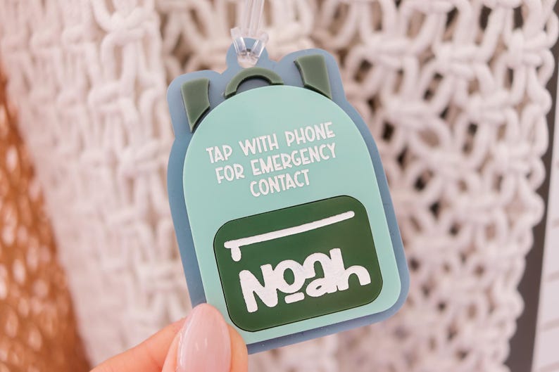 May include: A light blue backpack-shaped tag with the name "Noah" in white. The tag has the text "TAP WITH PHONE FOR EMERGENCY CONTACT". The tag is held up by a clear plastic loop.