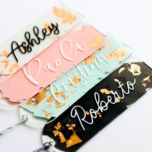 May include: Four personalized acrylic bookmarks with gold leaf accents. The bookmarks are in pink, mint green, and black with white lettering. The names on the bookmarks are Ashley, Pio-Ci, Christin, and Roberto.