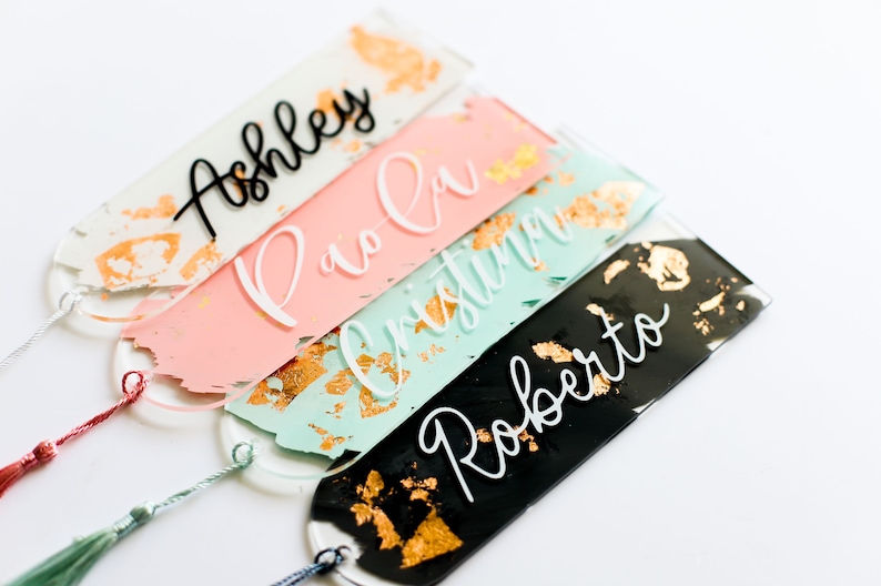 May include: Four personalized acrylic bookmarks with gold leaf accents. The bookmarks are in pink, mint green, and black with white lettering. The names on the bookmarks are Ashley, Pio-Ci, Christin, and Roberto.