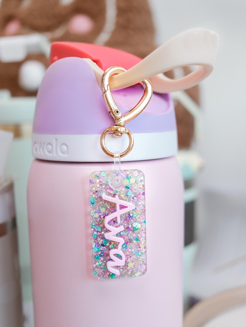 May include: A pink and purple water bottle with a gold-colored heart-shaped clasp and a glittery name tag that reads "Ava" in pink script. The bottle has a light pink body, a purple top, and a red flip-top lid.