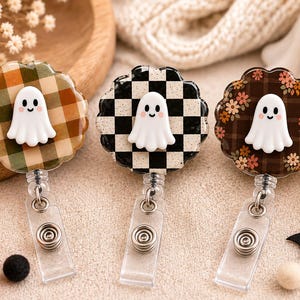 May include: Three circular badge reels featuring a white ghost figure on the front. The left reel has a green and orange plaid pattern, the center reel has a black and white checkerboard pattern, and the right reel has a brown floral plaid pattern.