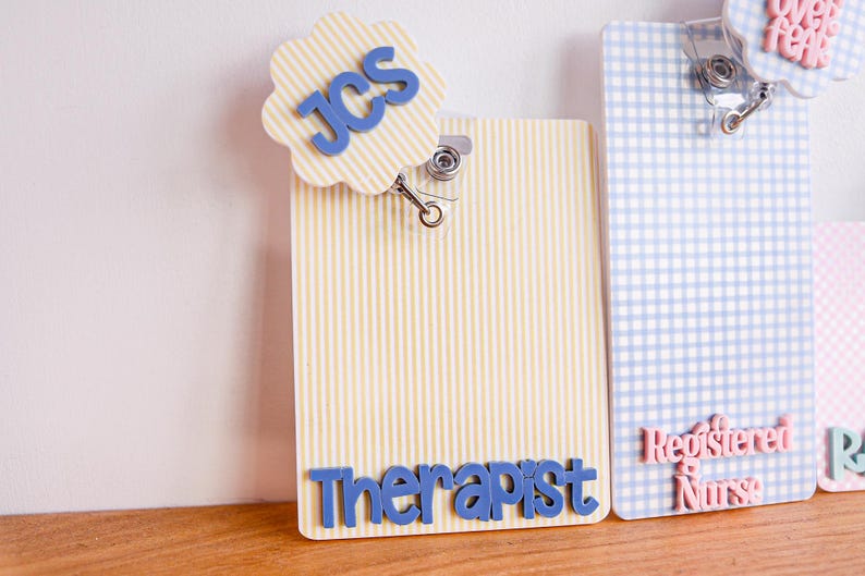 Puede incluir: Un porta credenciales de rayas verticales amarillas y blancas con un remate festoneado que presenta las letras azules JCS y el texto azul Therapist en la parte inferior. Al lado hay un porta credenciales de cuadros azules y blancos con texto rosa Registered Nurse.