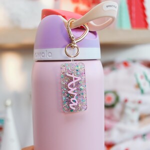 May include: A pink insulated water bottle with a purple lid and a gold-colored clip. A clear rectangular keychain with the name "Ava" in pink glitter is attached to the bottle.