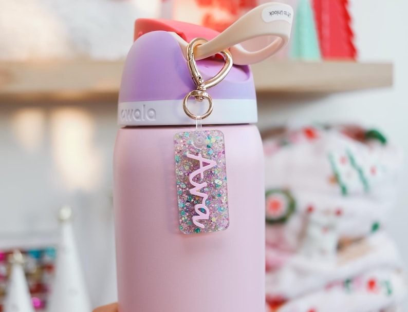 May include: A pink insulated water bottle with a purple lid and a gold-colored clip. A clear rectangular keychain with the name "Ava" in pink glitter is attached to the bottle.