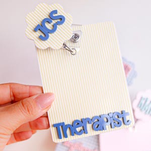 Puede incluir: Un porta credenciales rectangular con un patr&oacute;n de rayas verticales amarillas y blancas. Presenta un adorno circular festoneado con las letras azules JCS y la palabra Therapist en letras azules en la parte inferior, unido a un clip met&aacute;lico.