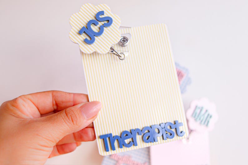 Puede incluir: Un porta credenciales rectangular con un patr&oacute;n de rayas verticales amarillas y blancas. Presenta un adorno circular festoneado con las letras azules JCS y la palabra Therapist en letras azules en la parte inferior, unido a un clip met&aacute;lico.