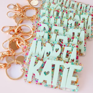 May include: A collection of personalized keychains with gold-colored hardware. The keychains feature names in a light green font with heart cutouts, set against a clear background filled with colorful glitter. The background is a soft pink.