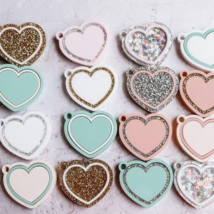 May include: A collection of 14 heart-shaped keychains with various designs. Some keychains are solid colors, while others feature glitter or a sparkly finish. The keychains are arranged in a semi-circle on a light gray background.