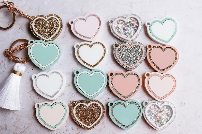 May include: A collection of 14 heart-shaped keychains with various designs. Some keychains are solid colors, while others feature glitter or a sparkly finish. The keychains are arranged in a semi-circle on a light gray background.