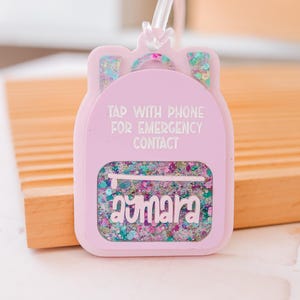 Puede incluir: Una etiqueta rosa claro con forma de mochila con el texto "TAP WITH PHONE FOR EMERGENCY CONTACT". La etiqueta tiene una secci&oacute;n transparente llena de purpurina colorida y el nombre "Aymara".