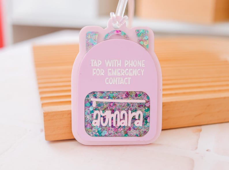 Puede incluir: Una etiqueta rosa claro con forma de mochila con el texto "TAP WITH PHONE FOR EMERGENCY CONTACT". La etiqueta tiene una secci&oacute;n transparente llena de purpurina colorida y el nombre "Aymara".