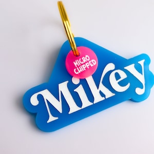 Pu&ograve; includere: Un'etichetta di identificazione per animali domestici in plastica blu con il nome "Mikey" in lettere bianche. Un cerchio rosa con il testo "MICRO CHIPPED" &egrave; attaccato all'etichetta.