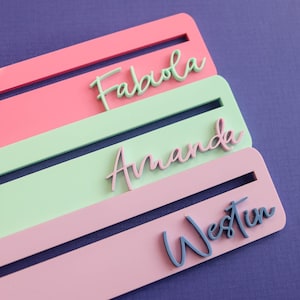 May include: Three rectangular acrylic strips in pink, light green, and light pink, each featuring a horizontal slot and a raised, cursive name cutout. The names are Fabiola in light green, Amanda in dark pink, and Westin in dark blue.