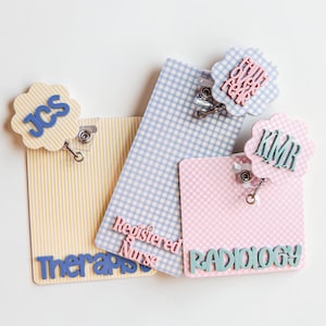 Puede incluir: Tres portatarjetas rectangulares con fondos estampados y remates festoneados. Uno de rayas amarillas dice JCS y Therapist. Uno de cuadros azules dice Registered Nurse. Uno de cuadros rosas dice KMR, Radiology y la frase Faith Over Fear.