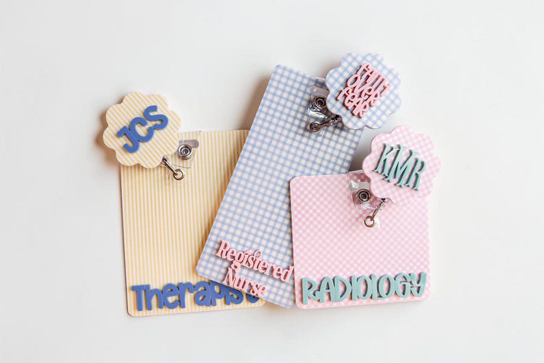 Puede incluir: Tres portatarjetas rectangulares con fondos estampados y remates festoneados. Uno de rayas amarillas dice JCS y Therapist. Uno de cuadros azules dice Registered Nurse. Uno de cuadros rosas dice KMR, Radiology y la frase Faith Over Fear.