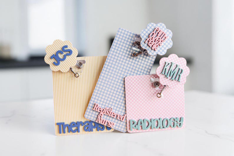 Puede incluir: Tres portatarjetas rectangulares con clips met&aacute;licos y adornos festoneados. Uno amarillo a rayas con JCS, uno azul a cuadros con Registered Nurse y uno rosa a cuadros con RADIOLOGY y KMR. Un adorno incluye el texto Faith Over Fear.