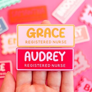 May include: Two name badges, one pink and one yellow, with the names "Grace" and "Audrey" in white and yellow lettering. Both badges read "Registered Nurse" in a smaller font. Other name badges are scattered in the background.
