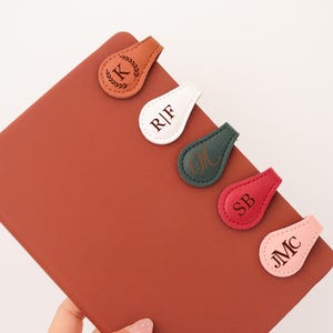 May include: A collection of personalized leather bookmarks in various colors, including tan, white, green, red, and pink. Each bookmark features initials and a unique design, resting on a brown notebook. The bookmarks are teardrop-shaped.