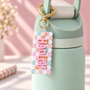 May include: A rectangular keychain with a scalloped edge featuring a checkered pattern in pastel pink, orange, and green. The name HADLEY is embossed in pink letters on the center. It is attached to a gold heart-shaped clasp on a sage green bottle.