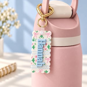 May include: A rectangular scalloped keychain with a white background featuring a pattern of pink bows, green leaves, and small pink flowers. The name Hannah is embossed in light blue letters. It is attached to a gold heart-shaped metal clasp.
