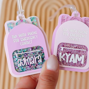 May include: Two light purple backpack-shaped luggage tags with glitter accents. Each tag has a clear strap and the text "TAP WITH PHONE FOR EMERGENCY CONTACT". One tag says "aymara" and the other says "KYAM".