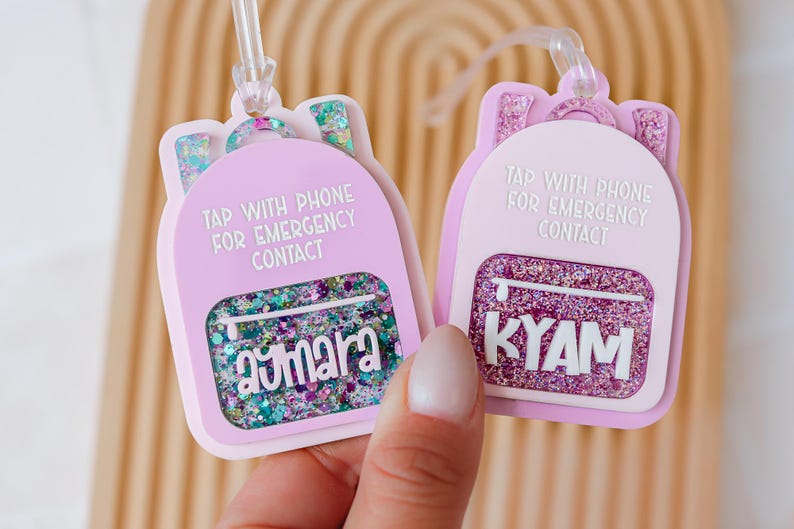 May include: Two light purple backpack-shaped luggage tags with glitter accents. Each tag has a clear strap and the text "TAP WITH PHONE FOR EMERGENCY CONTACT". One tag says "aymara" and the other says "KYAM".