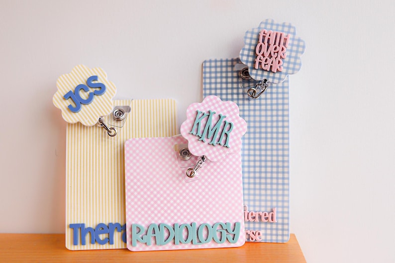 Puede incluir: Tres portatarjetas rectangulares con adornos circulares festoneados. Uno amarillo a rayas con JCS azul, uno rosa a cuadros con KMR y RADIOLOGY en azul verdoso, y uno azul a cuadros con el texto rosa FAITH OVER FEAR. Cada uno incluye cord&oacute;n retr&aacute;ctil.
