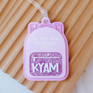 May include: A light pink backpack-shaped luggage tag with the text "TAP WITH PHONE FOR EMERGENCY CONTACT" and the name "KYAM". The tag has a glittery design and a clear plastic loop for attachment.