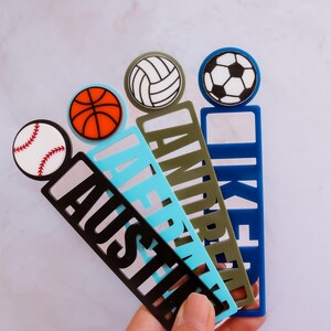 May include: Four colorful bookmarks with different sports ball designs. The bookmarks are made of acrylic and have the names "Austin", "LaFrentz", "Andrew", and "Iker" printed on them.
