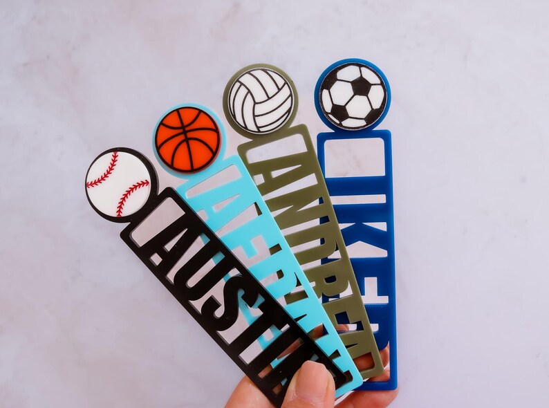 May include: Four colorful bookmarks with different sports ball designs. The bookmarks are made of acrylic and have the names "Austin", "LaFrentz", "Andrew", and "Iker" printed on them.