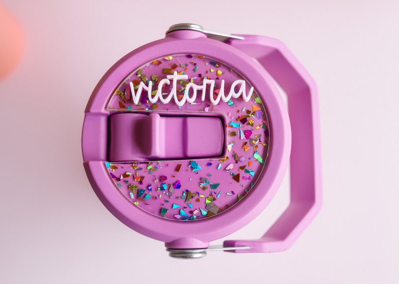 May include: A close-up of a lilac-colored water bottle lid with a handle. The lid features the name "Victoria" in white script and is filled with colorful glitter. The lid is made of plastic and has a flip-top closure.