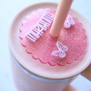 May include: A pink mug with a matching lid featuring a glittery, scalloped design. The lid has the word "mermaid" in white lettering, along with two butterfly accents. A pink straw extends from the lid.
