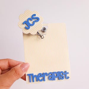 Puede incluir: Un portatarjetas rectangular con un patr&oacute;n de rayas verticales en color crema y blanco. La tarjeta presenta un adorno circular festoneado con el texto azul JCS y el texto azul Therapist en la parte inferior. Un clip met&aacute;lico sujeta el adorno.