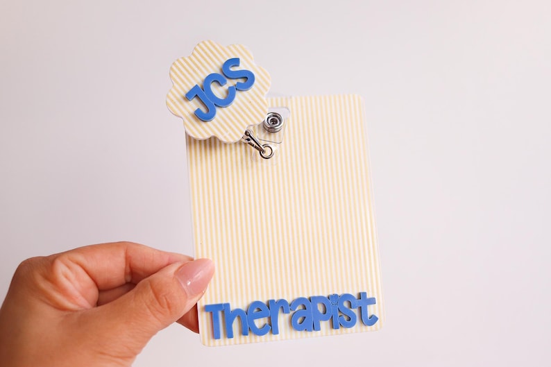 Puede incluir: Un portatarjetas rectangular con un patr&oacute;n de rayas verticales en color crema y blanco. La tarjeta presenta un adorno circular festoneado con el texto azul JCS y el texto azul Therapist en la parte inferior. Un clip met&aacute;lico sujeta el adorno.