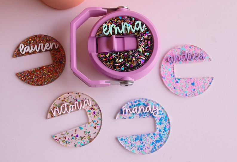 May include: A light purple water bottle with a handle and a glittery design. The bottle has the name "emma" on it. There are also several glittery name tags that say "lauren", "andrea", "victoria", and "mandy".