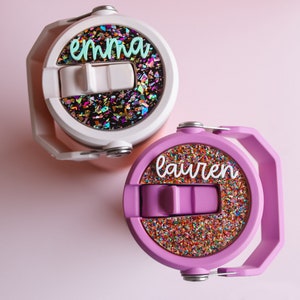 May include: Two personalized water bottles. One is white with the name "emma" in teal script, and the other is purple with the name "lauren" in white script. Both have a colorful glitter design on the lid.