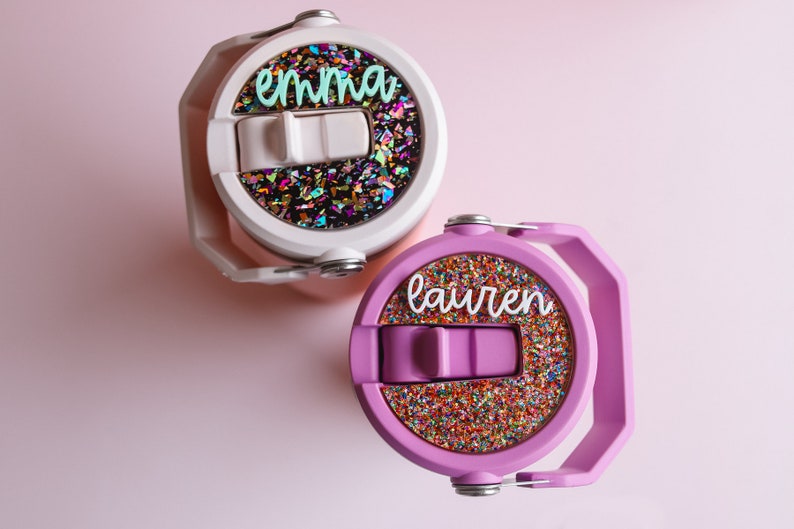 May include: Two personalized water bottles. One is white with the name "emma" in teal script, and the other is purple with the name "lauren" in white script. Both have a colorful glitter design on the lid.