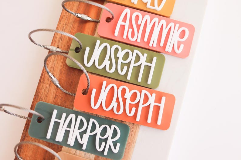 以下が含まれることがあります： 「Asmme」「Joseph」「Harper」などの白い名前が書かれた、カラフルなラゲッジタグのコレクション。長方形のタグは角が丸く、銀色のワイヤーのループが付いています。背景は木製のボードです。