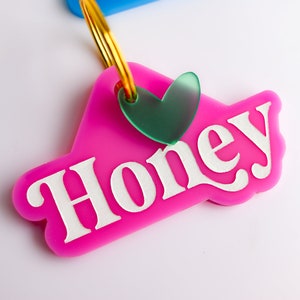 Pu&ograve; includere: Portachiavi in plastica rosa con la parola "Honey" in lettere bianche e un ciondolo a forma di cuore verde.