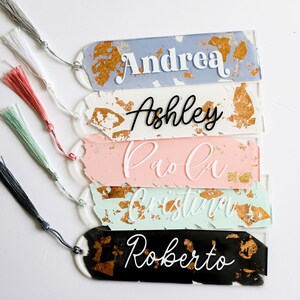 May include: Five personalized bookmarks with gold accents and tassels. The bookmarks are made of white, blue, pink, green, and black resin. The names Andrea, Ashley, Paola, Cristina, and Roberto are written on the bookmarks.