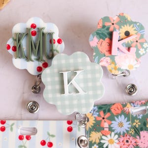 May include: Three floral-shaped badge reels with personalized initials. One features cherries and the initials "KMU", another has a floral pattern with the letter "K", and the third has a gingham pattern with the letter "K".