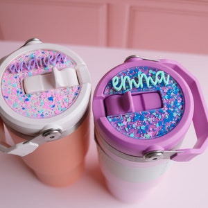 May include: Two personalized tumblers with lids. One is peach-colored with a white lid and the name "andrea" in purple. The other is purple with a light purple lid and the name "emma" in teal. Both lids have a glittery design.