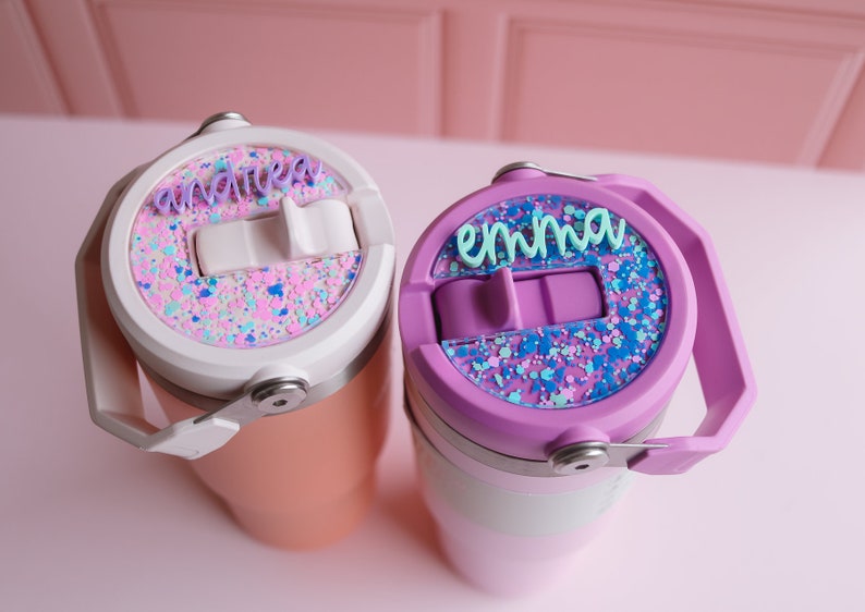 May include: Two personalized tumblers with lids. One is peach-colored with a white lid and the name "andrea" in purple. The other is purple with a light purple lid and the name "emma" in teal. Both lids have a glittery design.