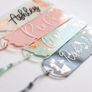 May include: Four personalized bookmarks with gold accents and tassels. The bookmarks are made of clear acrylic and have a painted design. The names "Ashley", "Paola", "Christina", and "Roberto" are written on the bookmarks in white and black lettering.