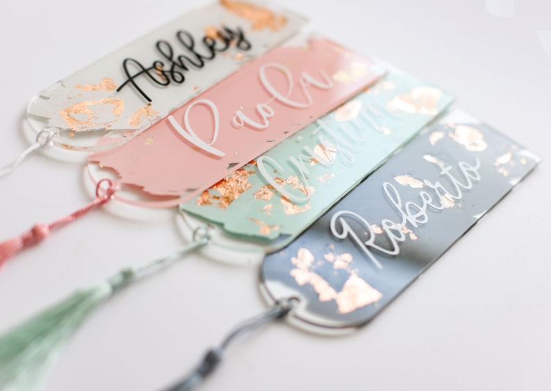 May include: Four personalized bookmarks with gold accents and tassels. The bookmarks are made of clear acrylic and have a painted design. The names "Ashley", "Paola", "Christina", and "Roberto" are written on the bookmarks in white and black lettering.